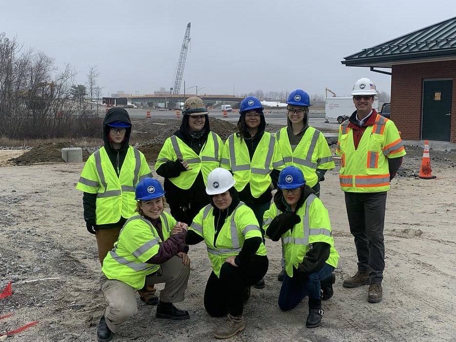 a group of young students huddle together, both standing and kneeling, for a group photo with CPM Constructors CFO/CEO, Tim Ouellette, with the CPM Constructions Exit 45 project in the background. 